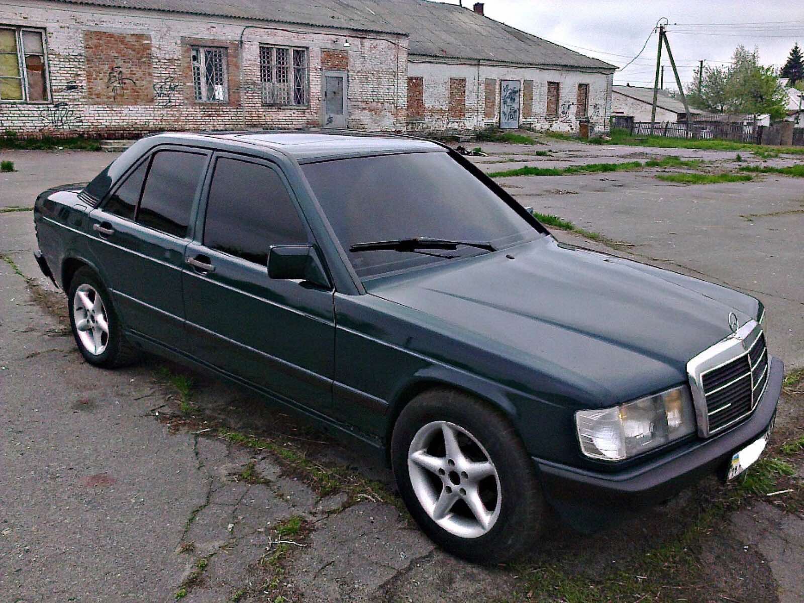 Ciemnozielony Mercedes-Benz W201 z pięcioramiennymi felgami, zaparkowany na betonowym placu przed ceglanym budynkiem z zabitymi oknami.