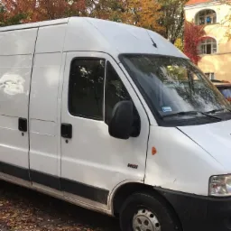 Biały samochód dostawczy typu furgon, zaparkowany na brukowanym parkingu z opadłymi liśćmi, na tle budynku z czerwoną winoroślą i jesiennych drzew.