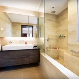 Wykończona łazienka z beżowymi płytkami imitującymi marmur, dużą umywalką na szafce z ciemnego drewna i kabiną prysznicową oddzieloną szybą.