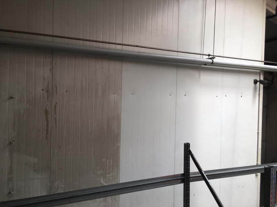 Fragment przemysłowej ściany panelowej w trakcie czyszczenia, widoczna wyraźna różnica między brudną a umytą powierzchnią, z rurami instalacyjnymi biegnącymi wzdłuż ściany.