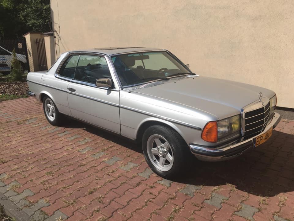 Srebrny Mercedes-Benz W123 Coupe na tle jasnej ściany i kostki brukowej, widok z przodu lekko z boku, z widoczną tablicą rejestracyjną.