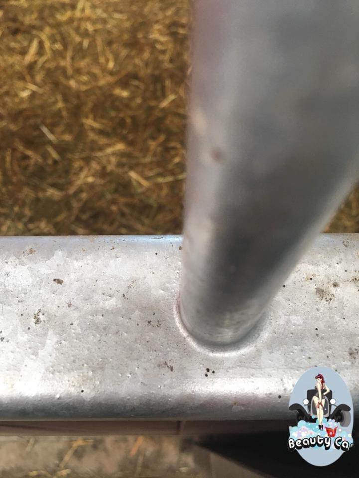 Detail of a dirty, galvanized metal structure, possibly a barrier or railing, with straw or hay visible in the background and a 'Beauty Car' logo sticker affixed.