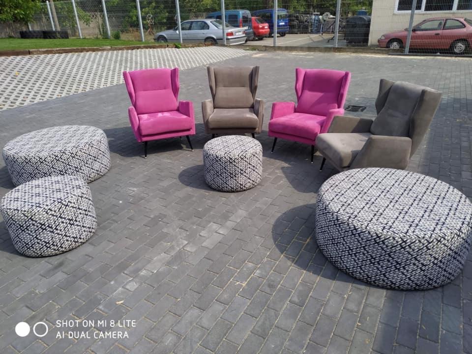 Dwa fotele w odcieniach różu i dwa w odcieniach szarości, ustawione na kostce brukowej wraz z pięcioma okrągłymi pufami w biało-szarą kratę, na tle zaparkowanych samochodów i ogrodzenia.