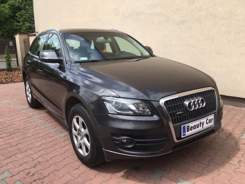 Ciemnoszary SUV marki Audi Q5 zaparkowany na kostce brukowej przed jasną ścianą budynku, z tabliczką 'Beauty Car' na przednim zderzaku.