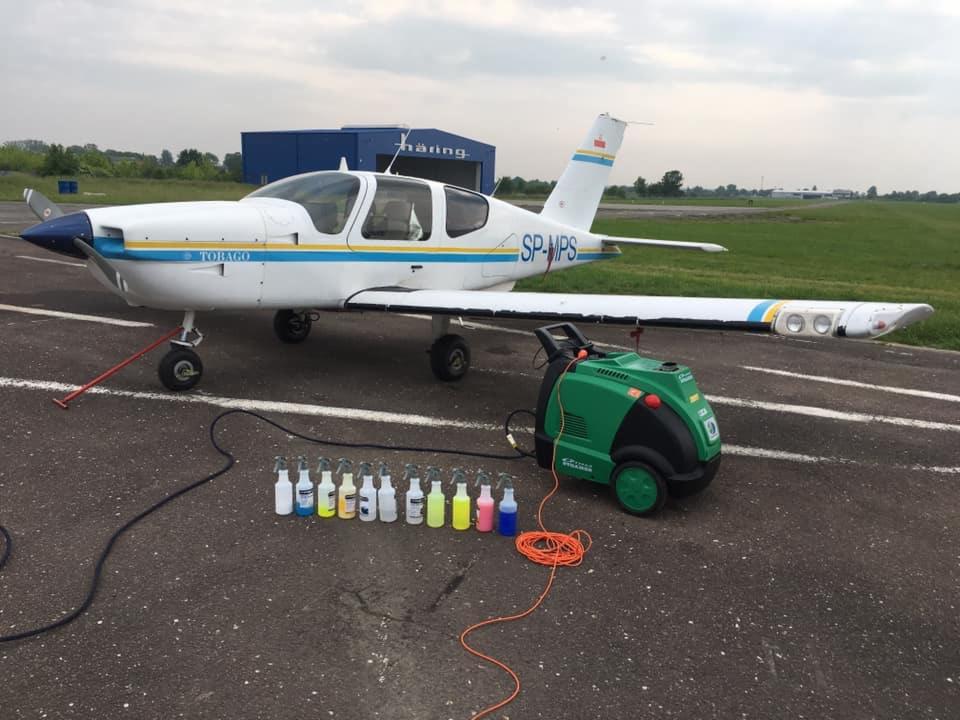 Samolot Tobago SP-MPS na lotnisku, obok niego zielona myjka ciśnieniowa i rząd kolorowych butelek z detergentami.