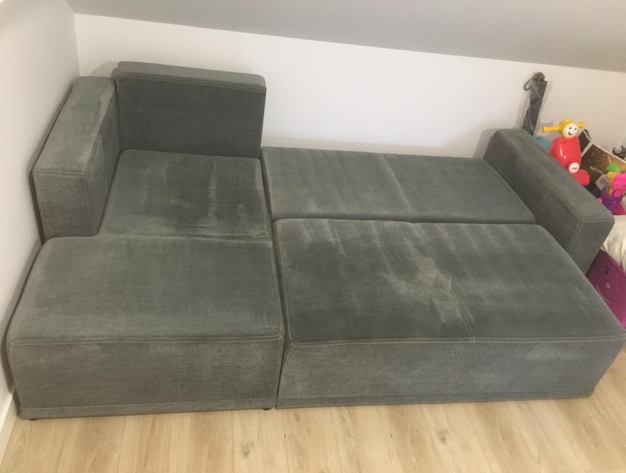Rozłożona szara kanapa z tkaniny w pokoju z zabawkami w tle, widoczne ślady użytkowania na siedzisku.