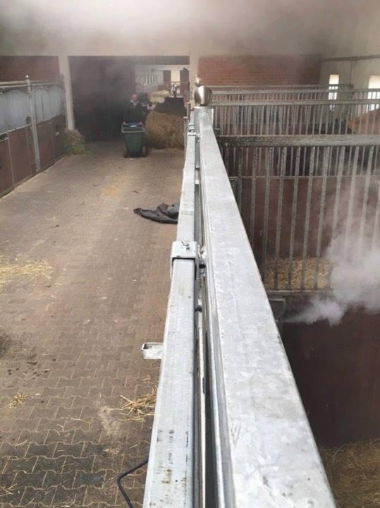 Wnętrze stajni z widocznymi boksami dla koni, gdzie z jednego z boksów wydobywa się para, prawdopodobnie po czyszczeniu parowym. W tle widoczna osoba z wózkiem i stertą siana.