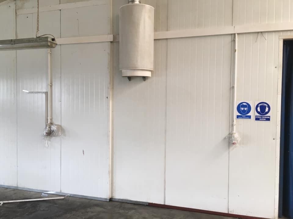 Wnętrze hali produkcyjnej z panelami ściennymi, oświetleniem i znakiem BHP nakazującym stosowanie ochrony oczu i słuchu.