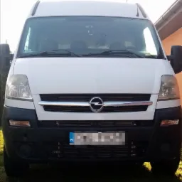 Biały Opel Movano, widok z przodu, z zamazaną tablicą rejestracyjną na tle trawnika i budynku.