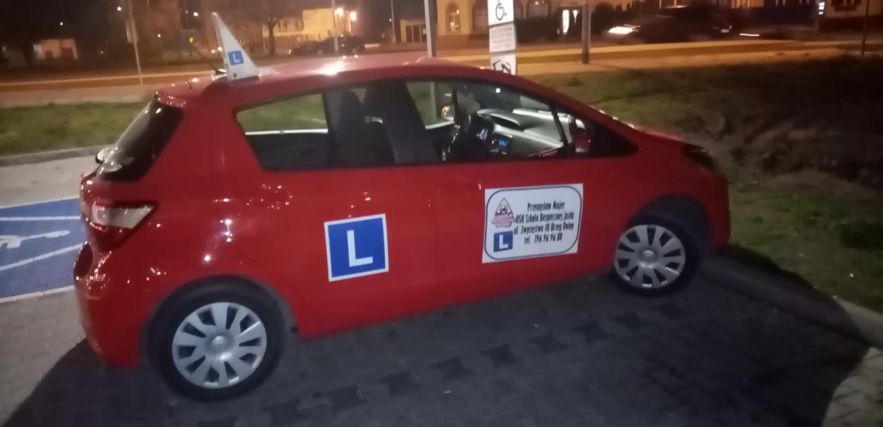 Czerwony samochód z literą L, stojący na parkingu z wyznaczonym miejscem dla osób niepełnosprawnych, nocne oświetlenie uliczne.