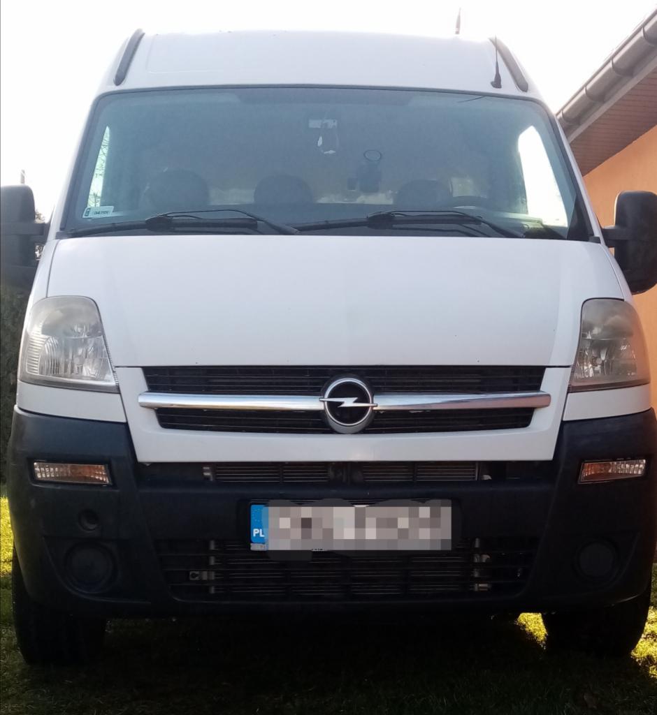 Biały Opel Movano, widok z przodu, z zamazaną tablicą rejestracyjną na tle trawnika i budynku.