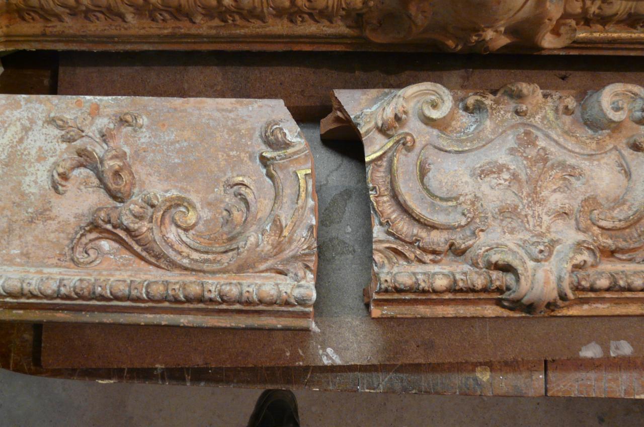 Fragmenty zabytkowej, zdobionej sztukaterii w odcieniach beżu i złota, z widocznymi uszkodzeniami i śladami wcześniejszych napraw, ułożone obok siebie na drewnianym podłożu.