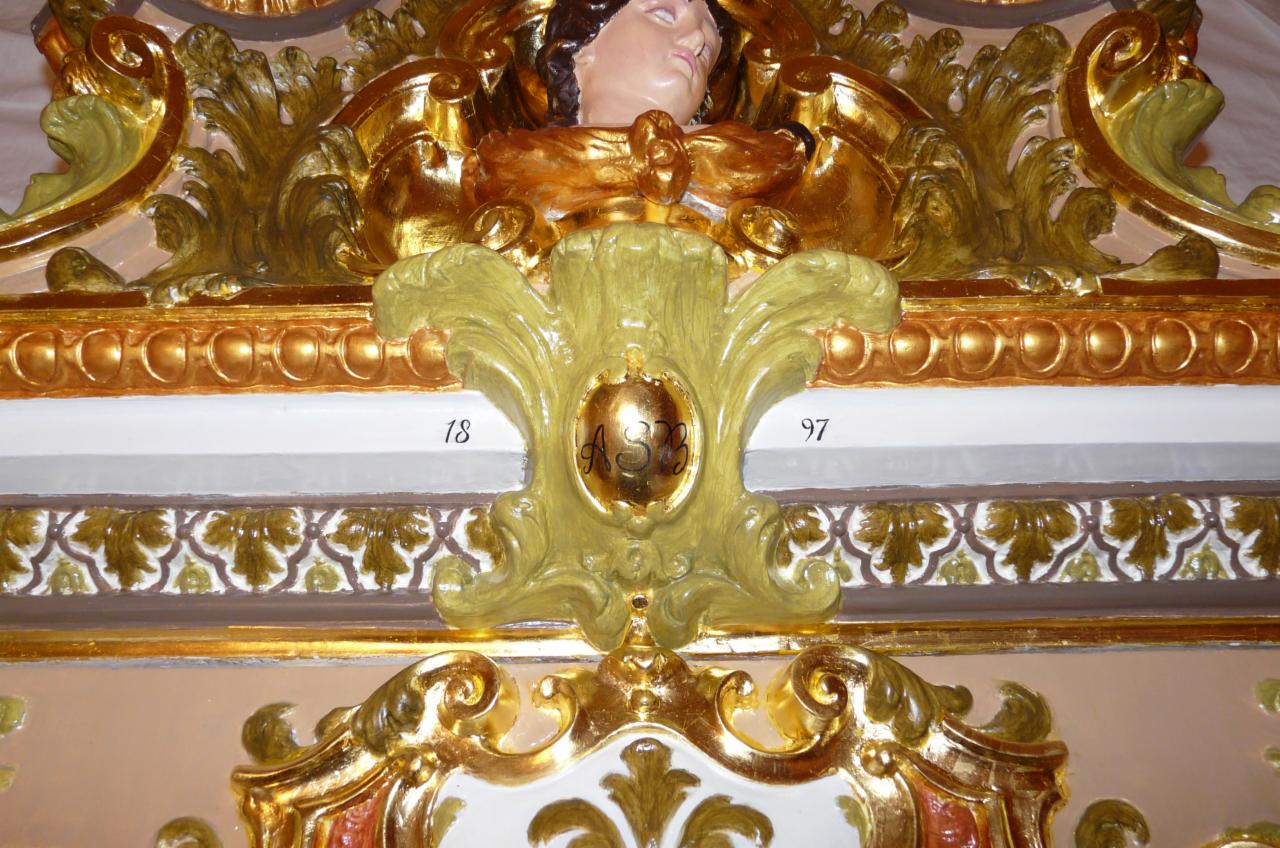 Fragment dekoracji ściennej z rzeźbionymi elementami, złoceniami i numerami '18' i '97' po bokach centralnego elementu z inicjałami 'ASB'.