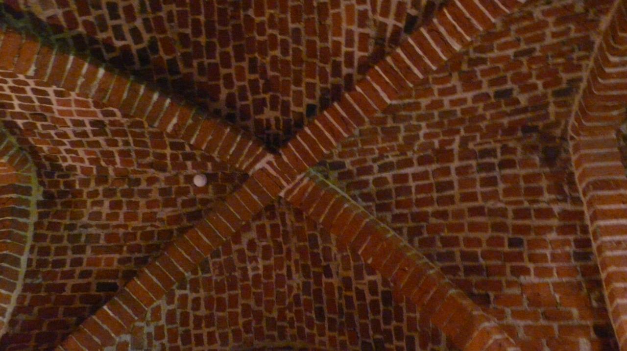 Sklepienie krzyżowo-żebrowe z czerwonej cegły, widok z dołu, z widocznymi śladami napraw i niewielkim punktowym źródłem światła