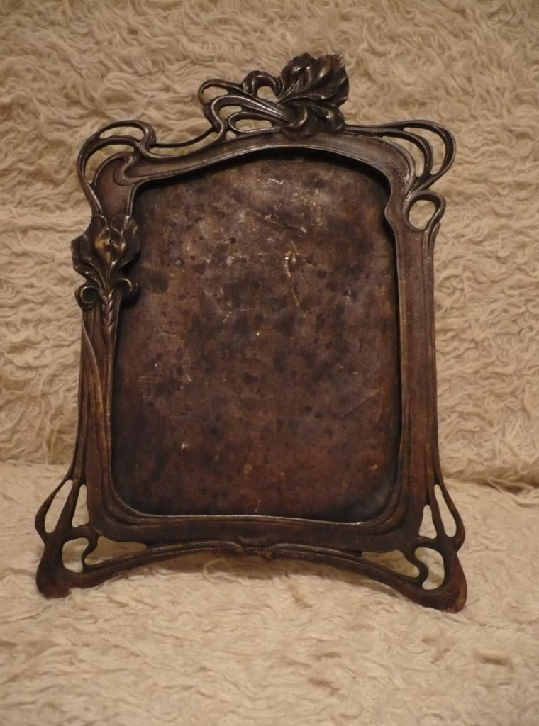 Metalowa, secesyjna ramka na zdjęcie z Elbląga, z motywami kwiatowymi i śladami patyny, ustawiona na teksturowanym tle.