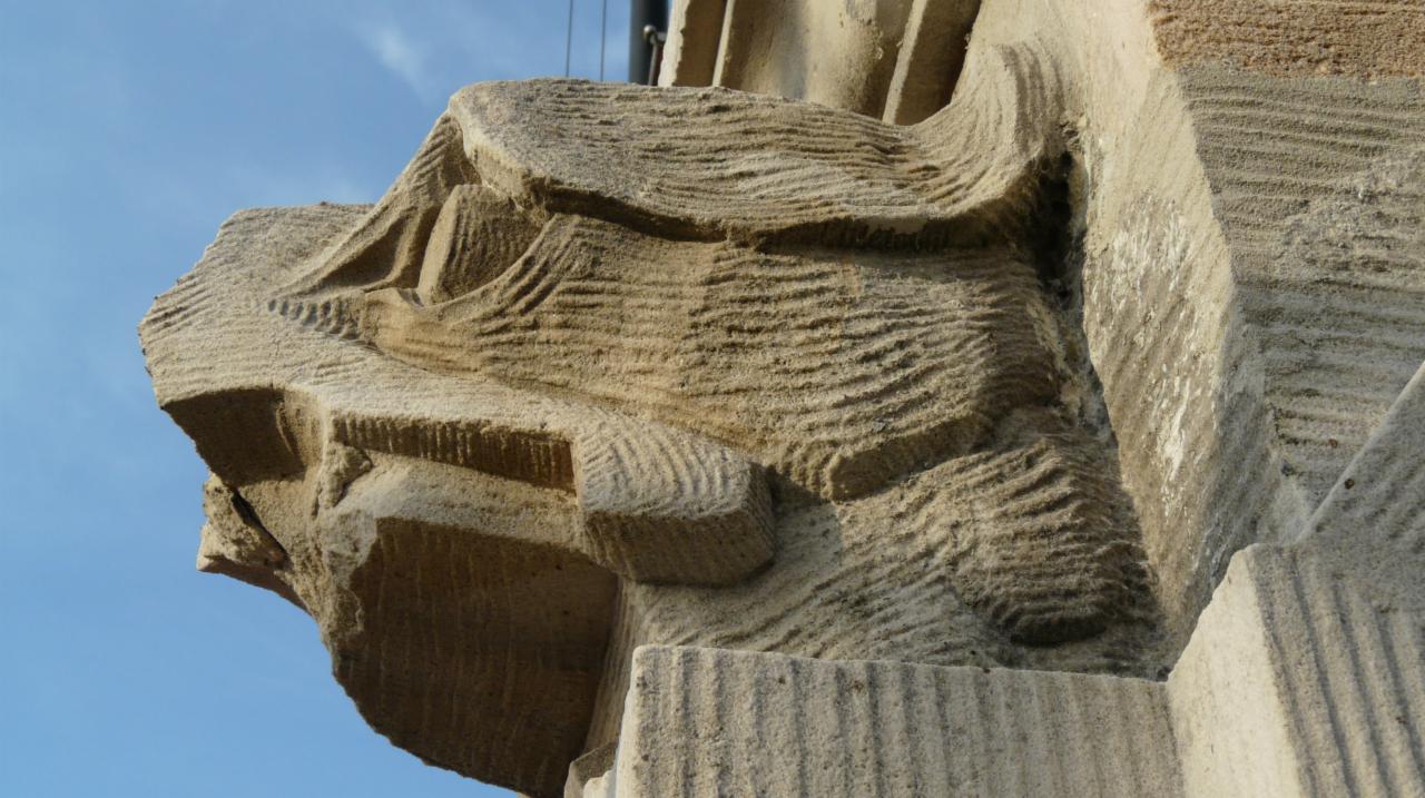 Detal architektoniczny z piaskowca z wyraźnymi śladami erozji i charakterystycznymi, falistymi wzorami obróbki kamienia, widoczny na tle błękitnego nieba.