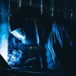 Spawacz w masce ochronnej, otoczony niebieskim dymem i iskrami podczas pracy.