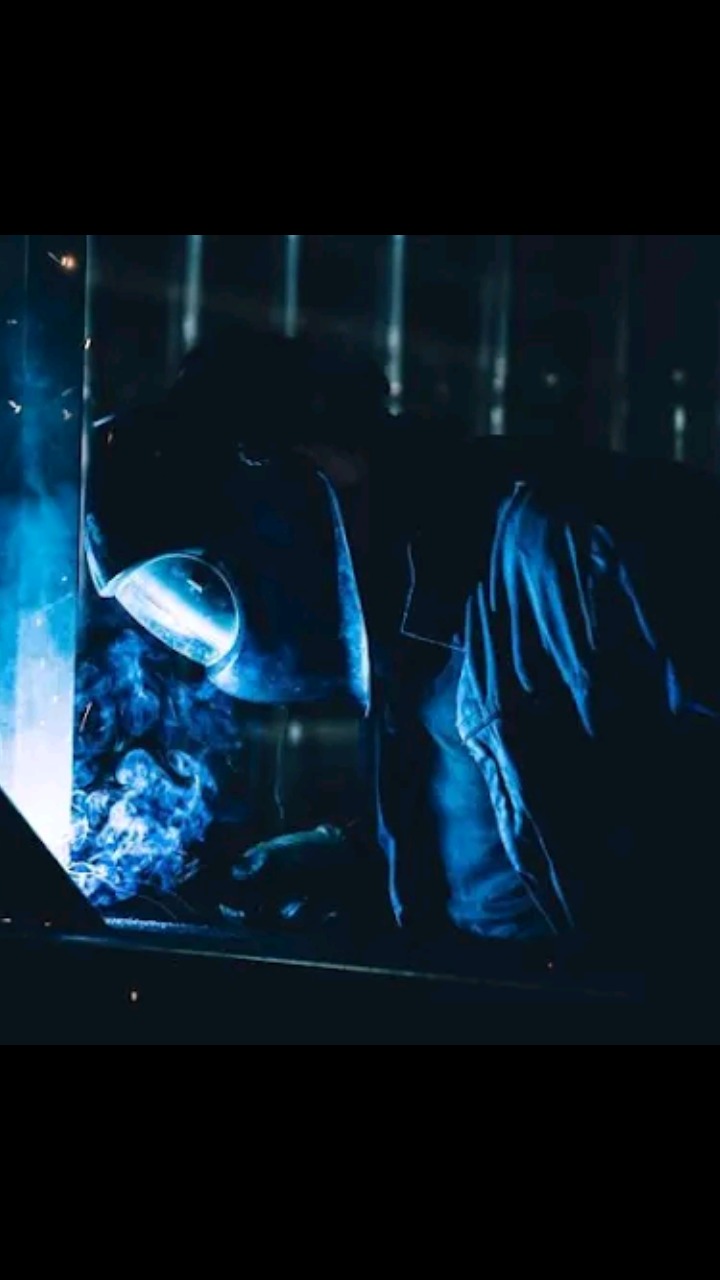 Spawacz w masce ochronnej, otoczony niebieskim dymem i iskrami podczas pracy.