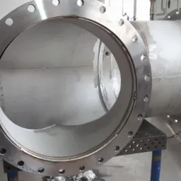 Element rurociągu ze stali nierdzewnej z przyspawanymi kołnierzami, widoczne spoiny i otwory montażowe, umieszczony na stole spawalniczym.