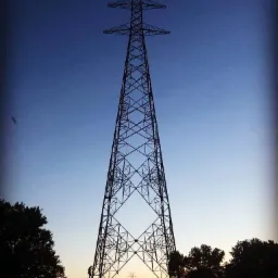 Sylwetka słupa wysokiego napięcia o konstrukcji kratownicowej na tle zachodzącego słońca, z pracownikiem wspinającym się po jego konstrukcji.