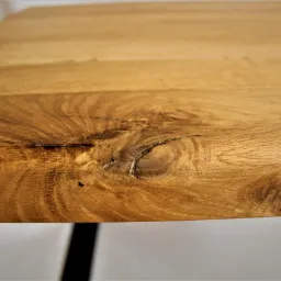 Zbliżenie na krawędź blatu z jasnego drewna z wyraźnym usłojeniem i sękiem, wspartego na czarnych, metalowych nogach.