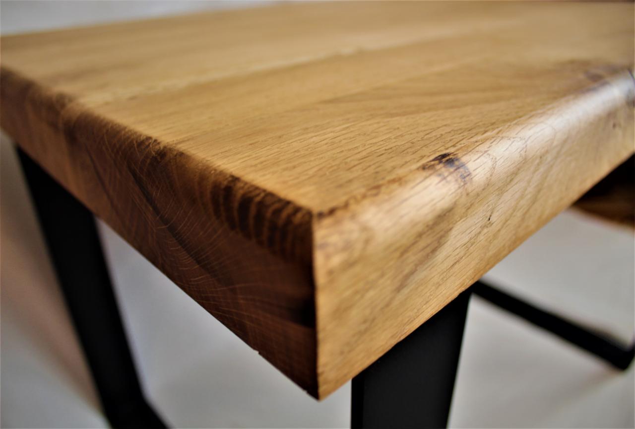 Krawędź blatu z jasnego drewna dębowego, z widocznym usłojeniem i zaokrąglonymi brzegami, osadzona na czarnej, metalowej podstawie w kształcie litery X.