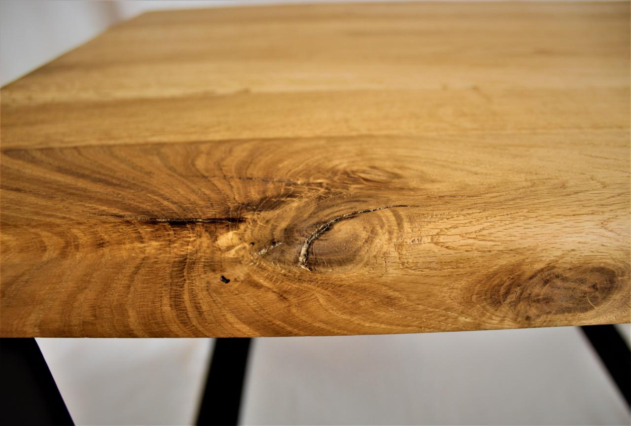 Zbliżenie na krawędź blatu z jasnego drewna z wyraźnym usłojeniem i sękiem, wspartego na czarnych, metalowych nogach.