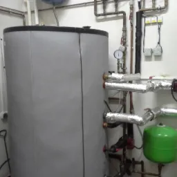 Szary zbiornik buforowy ciepła z rurami w izolacji aluminiowej, manometry i zielony zbiornik wyrównawczy w kotłowni w Gdańsku.