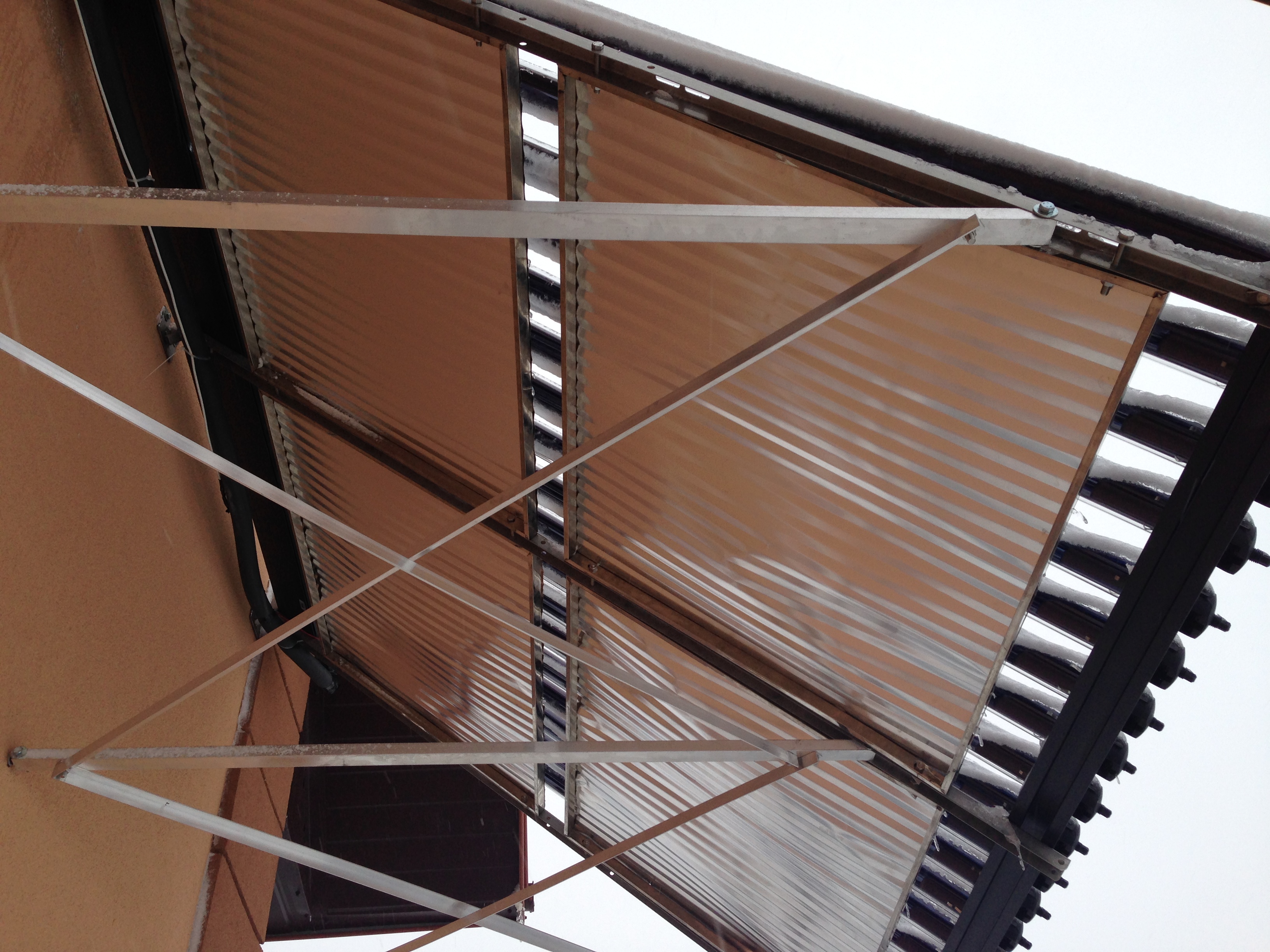 Szczegółowy widok od dołu na kolektory słoneczne z pokrywą z blachy falistej, oszronione śniegiem, zamontowane na metalowej konstrukcji wsporczej przy ścianie budynku.