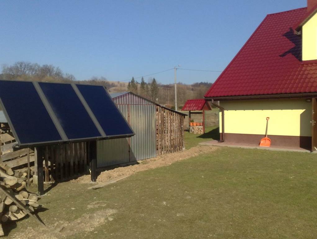 Panele słoneczne zamontowane na drewnianej konstrukcji przed budynkiem gospodarczym, w tle dom z czerwonym dachem i łopata oparta o ścianę.