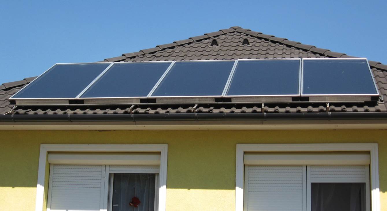 Panele słoneczne zamontowane na dachu domu z żółtą elewacją i brązową dachówką, widoczne białe rolety okienne.