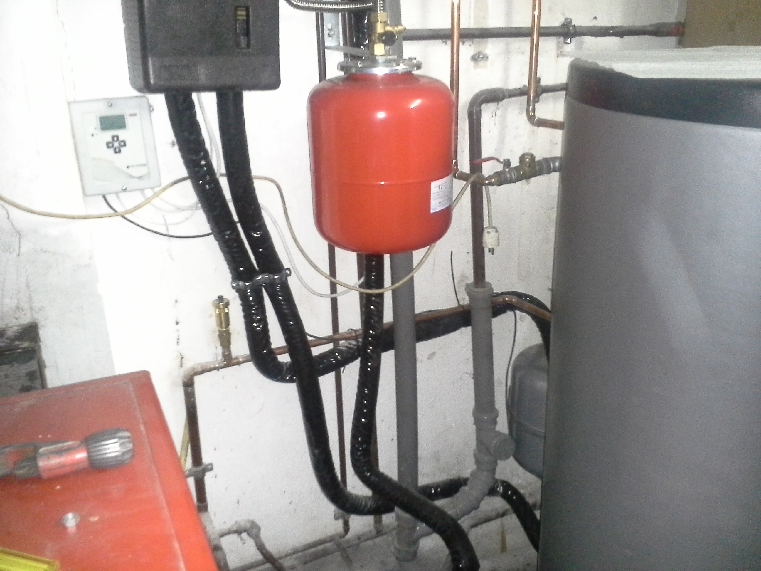 Czerwony zbiornik wyrównawczy z podłączonymi rurami w kotłowni, widoczny manometr i czujnik temperatury.