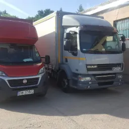 Dwa samochody dostawcze: czerwony Fiat Ducato z zabudową kontenerową i szary DAF LF z plandeką, zaparkowane obok budynku.