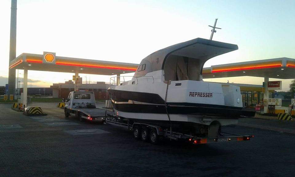 Łódź motorowa 'REPRESSER' na trzyosiowej przyczepie, transportowana przez małą lawetę na tle stacji benzynowej Shell.