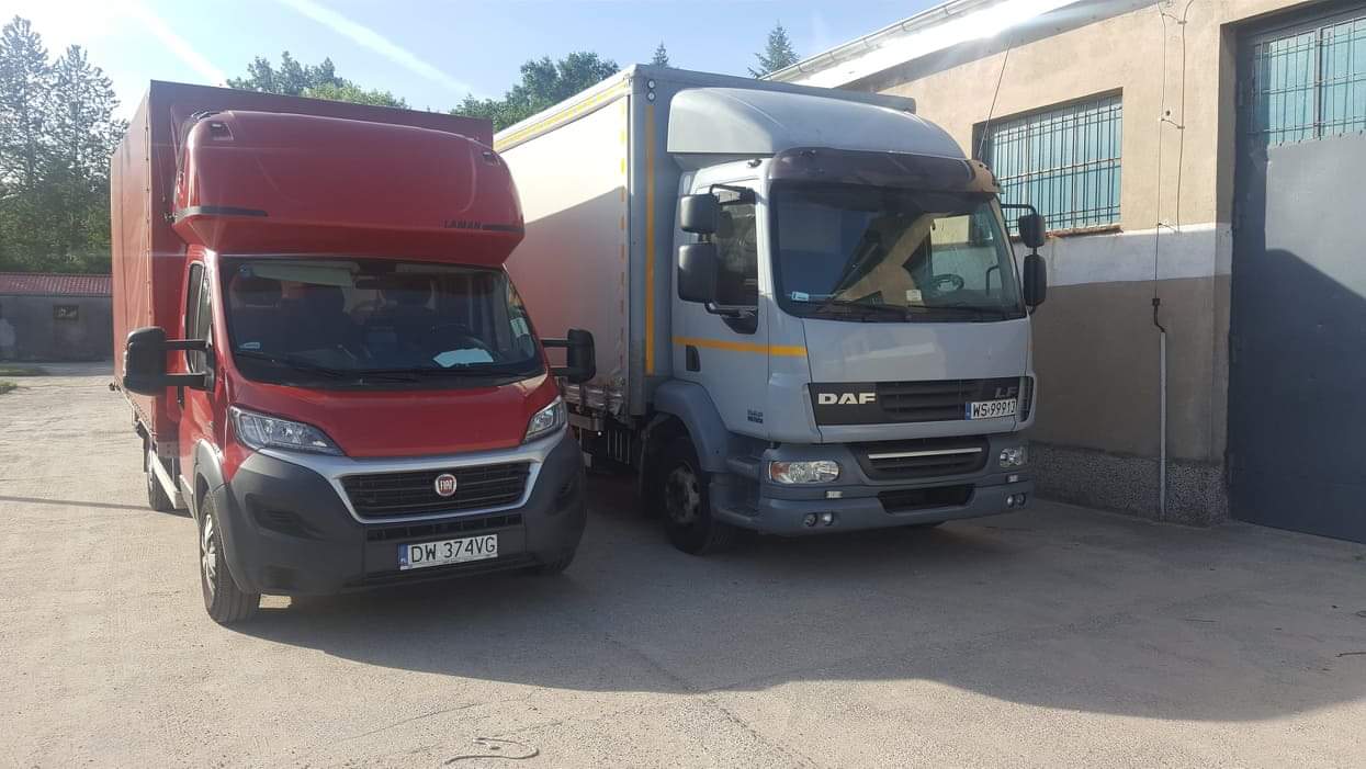 Dwa samochody dostawcze: czerwony Fiat Ducato z zabudową kontenerową i szary DAF LF z plandeką, zaparkowane obok budynku.