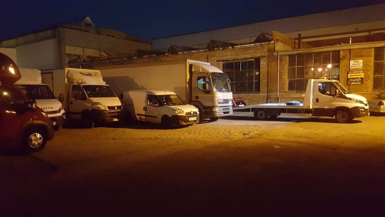 Flota pojazdów transportowych: busy Iveco, Fiat Doblo, ciężarówka DAF i laweta Iveco zaparkowane na placu w słabym oświetleniu miejskim.