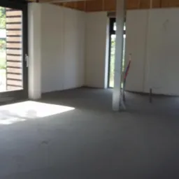 Surowe wnętrze w trakcie wykańczania: wylewka podłogowa, ściany z płyt kartonowo-gipsowych, duże okno przesuwne z widokiem na zieleń, widoczne instalacje i drewniane elementy konstrukcyjne...