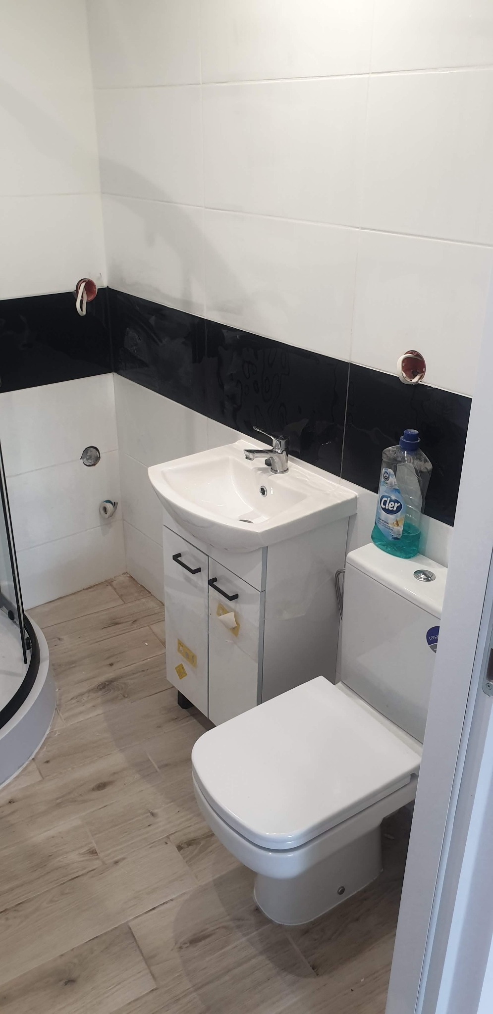 Wykończona łazienka z biało-czarnymi płytkami, umywalką z szafką, toaletą i drewnopodobną podłogą; widoczna butelka płynu do czyszczenia na spłuczce.