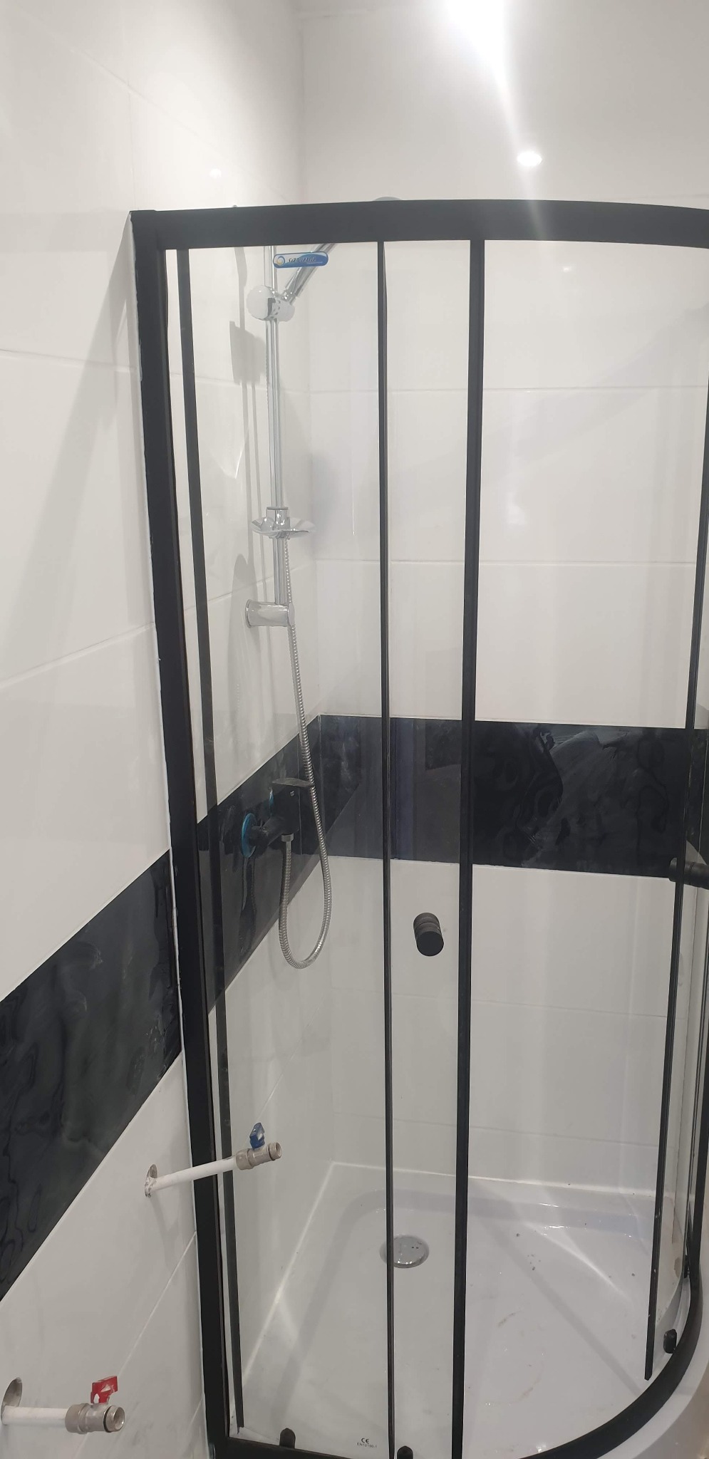 Nowoczesna kabina prysznicowa z czarną ramą i szklanymi drzwiami, zamontowana w łazience z białymi płytkami i pasem czarnego marmuru, widoczne przyłącza hydrauliczne.