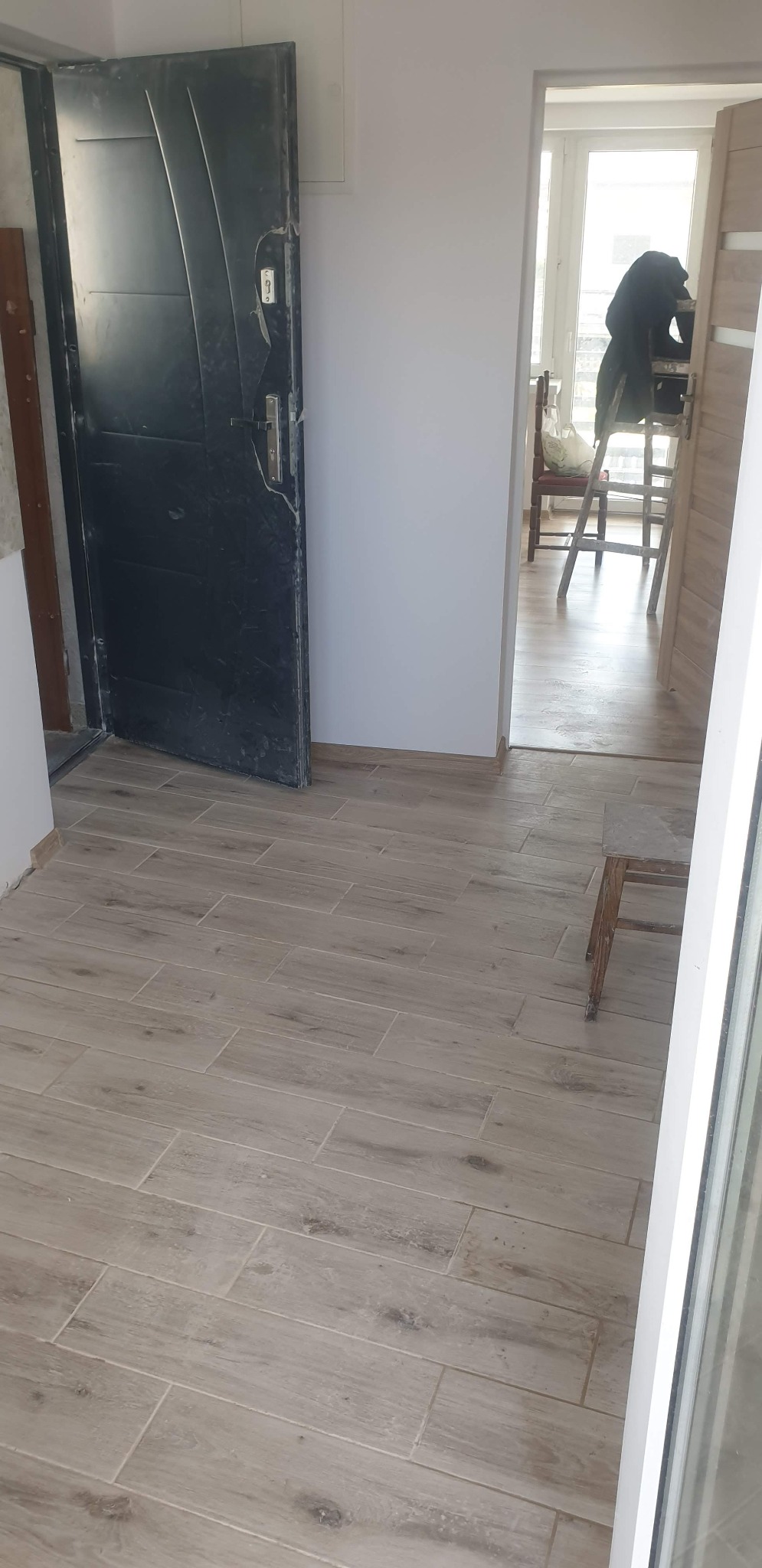 Ułożona podłoga z płytek imitujących drewno w jasnym odcieniu, widoczna boczna ściana i czarne drzwi wejściowe, w tle pomieszczenie z drabiną malarską.