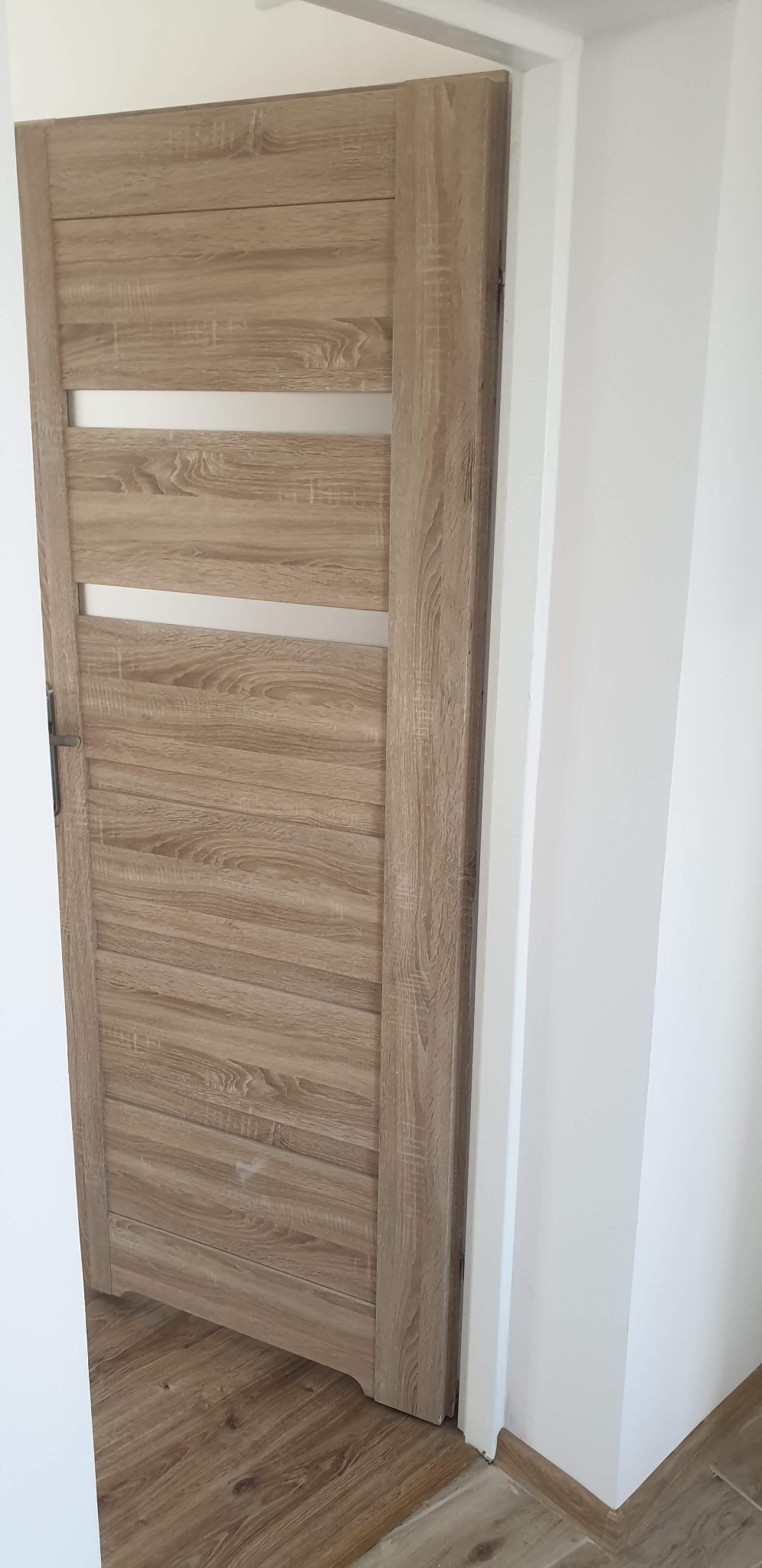 Zamknięte, drewnopodobne drzwi wewnętrzne z poziomymi, matowymi przeszkleniami, zamontowane w białej ościeżnicy, widoczne panele podłogowe.