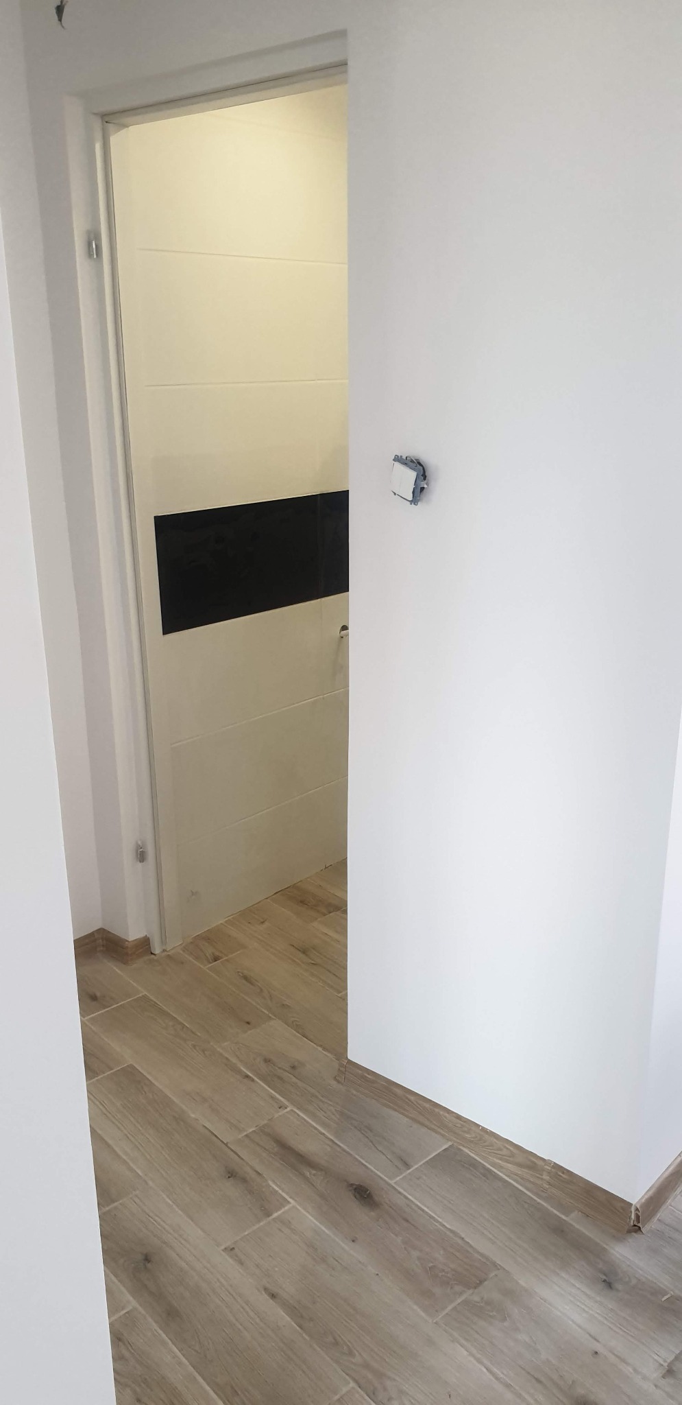 Widok na wykończone wnętrze: jasne płytki podłogowe imitujące drewno, białe ściany z zamontowanym gniazdkiem elektrycznym bez obudowy oraz drzwi z poziomym czarnym pasem dekoracyjnym.