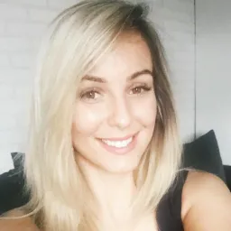 Uśmiechnięta blondynka w czarnej bluzce, z delikatnym makijażem, na tle białej ściany i ciemnej sofy. Portret w naturalnym świetle.