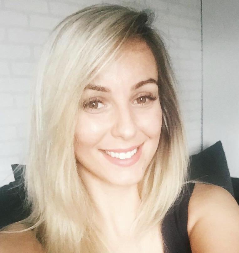 Uśmiechnięta blondynka w czarnej bluzce, z delikatnym makijażem, na tle białej ściany i ciemnej sofy. Portret w naturalnym świetle.