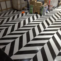 Ukończona podłoga z czarno-białych płytek ułożonych we wzór jodełki, widoczne narzędzia i materiały budowlane w tle.