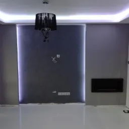 Wykończone wnętrze pokoju z szarymi ścianami, podświetlanym sufitem, błyszczącą podłogą i czarnym żyrandolem. Widoczne gniazdka elektryczne i wnęka w ścianie.