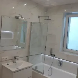 Elegancka łazienka w bieli i marmurze, z kabiną prysznicową nad wanną, umywalką z szafką i dużym lustrem podświetlonym od góry.