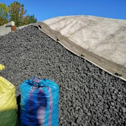 Góra czarnego, drobnego węgla kamiennego przykryta plandeką, z dwoma workami: żółtym i niebieskim, na tle błękitnego nieba.