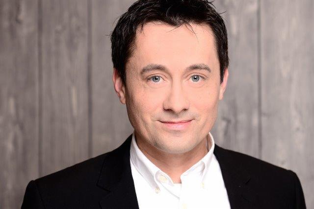 Portret mężczyzny w czarnej marynarce i białej koszuli na tle drewnianej ściany. Uśmiechnięty, z delikatnym makijażem. Profesjonalny wizerunek.