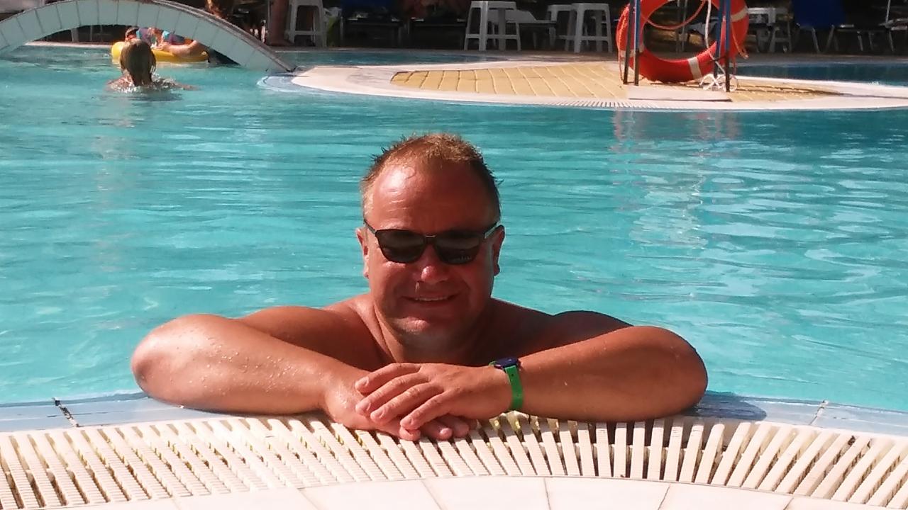 Uśmiechnięty mężczyzna w okularach przeciwsłonecznych opiera się o brzeg basenu z błękitną wodą. W tle widoczny mostek i osoby w pontonach.